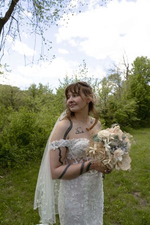 Bride in white lace strapless gown holds floral bouquet while gazing upward in a green outdoor setting.