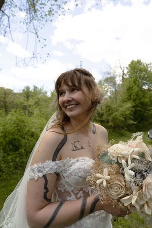 Smiling bride in off-shoulder lace gown holds bouquet outdoors under bright blue sky.