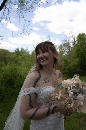 Smiling bride in off-shoulder lace gown and veil holding floral bouquet in a lush outdoor setting.