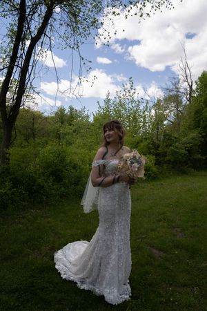 Bride in lace off-shoulder mermaid gown holding bouquet of neutral flowers in a lush green outdoor setting.