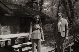 Black and white photo of couple in matching suits posing near rustic stone cabin at outdoor venue.