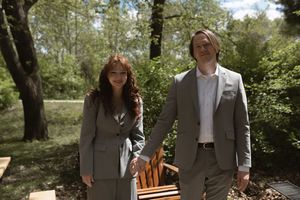 Couple in matching gray suits walking together in a sunlit outdoor garden setting.