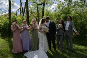 Bride and groom kissing surrounded by smiling wedding party in colorful dresses on green lawn.
