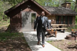 Dapper groom in dark suit walking confidently outside a rustic wooden cabin surrounded by trees.