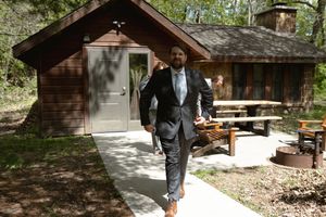 Well-dressed man in a dark suit walks confidently along a path outside a rustic wooden cabin in a forest setting.