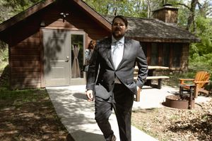 Groom in dark suit walking confidently along a path outside a rustic wooden cabin on wedding day.