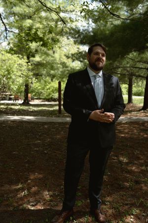 Handsome groom in dark suit and light tie stands confidently in a shaded woodland garden setting.