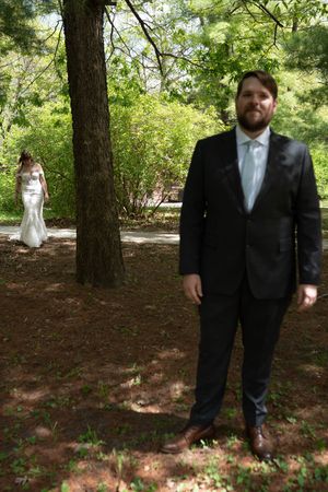 Groom in black suit stands in wooded park with bride in white lace gown visible in background among trees.