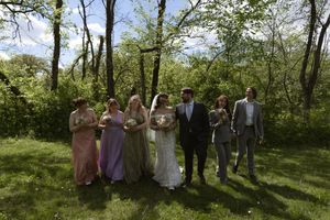 Wedding party poses in sunlit green meadow with bridesmaids in colorful gowns and groom in gray suit.