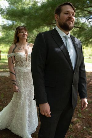 Groom in charcoal suit with mint tie stands outdoors as bride in lace gown approaches from behind.