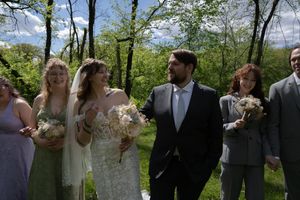 Wedding party walks outdoors in lush green setting, bride in white holding bouquet beside groom in gray suit.