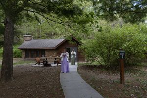 Two bridesmaids in purple and sage gowns stand near rustic cabin on wooded pathway at wedding venue.