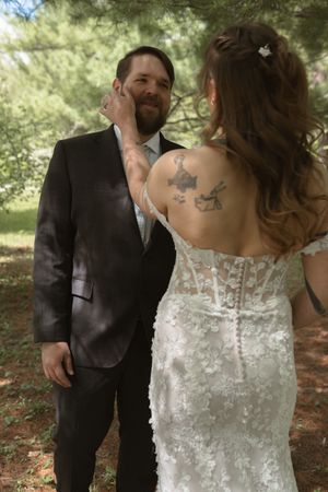 Bride with tattoo in lace wedding dress touching grooms face during first look among pine trees.