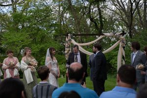 Bride and groom exchange vows under draped wooden arch surrounded by wedding party in lush green garden.