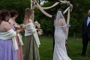 Bride in lace veil gown stands at floral arch with bridesmaids during outdoor wedding ceremony.