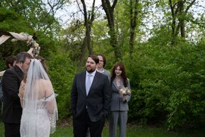 Groom in gray suit gazes at veiled bride during outdoor wedding ceremony beneath fabric-draped wooden arch.