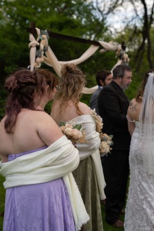Bridesmaids in pastel gowns with white shawls hold bouquets while standing at an outdoor wedding ceremony.