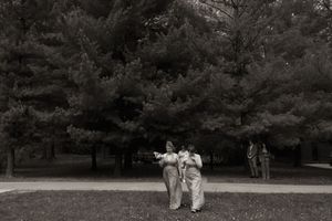 Black and white photo of two people in matching outfits embracing on grass near tall pine trees.