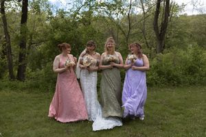 Bride poses with three bridesmaids in colorful pink, silver, and lavender gowns in a green outdoor setting.
