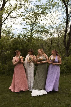 Bride with three bridesmaids in pink, sage, and lavender lace gowns holding bouquets outdoors.