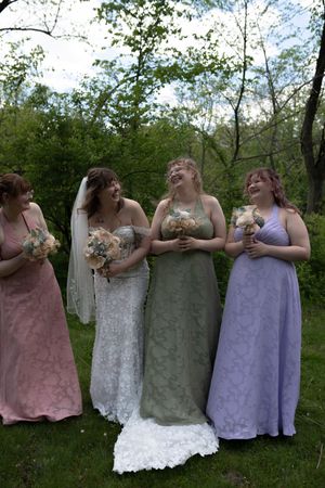 Bride with three bridesmaids in pink, green, and lavender gowns laughing outdoors.