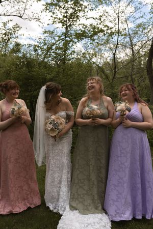 Bride in lace gown with three bridesmaids in pink, sage, and lavender dresses holding bouquets outdoors.