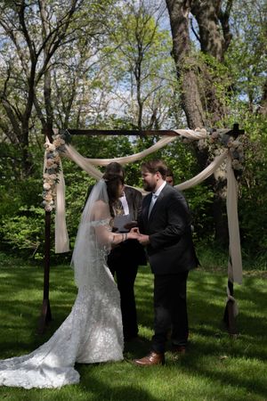 Bride and groom exchange vows under a rustic wooden arch adorned with fabric and flowers in a lush outdoor garden ceremony.