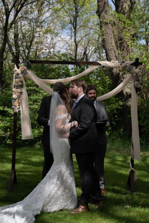 Bride and groom share first kiss under rustic wooden arch draped with fabric at outdoor garden wedding ceremony.