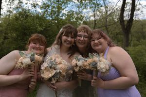 Bride with three bridesmaids in purple dresses holding bouquets of cream and sage flowers at outdoor wedding.