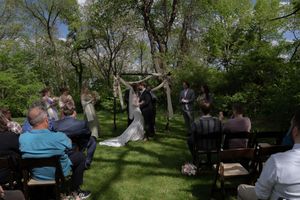 Outdoor wedding ceremony under rustic arch with guests seated among lush green trees.