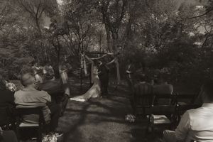 Black and white photo of an outdoor wedding ceremony with guests seated on wooden chairs among trees.