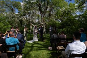 Outdoor wedding ceremony with guests seated on chairs as couple exchanges vows under a floral arch in a lush green garden.