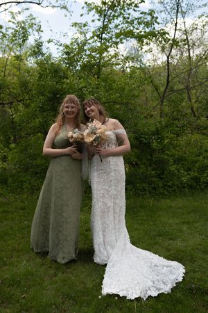Bride in white lace gown and bridesmaid in sage green dress pose outdoors with bouquet.