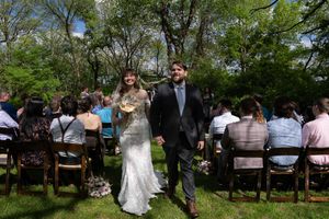 Newlyweds walk hand-in-hand down outdoor aisle after wedding ceremony surrounded by seated guests.