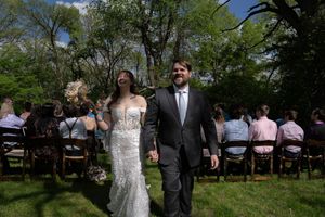 Newlyweds walk hand in hand down the aisle after outdoor wedding ceremony with seated guests and trees.