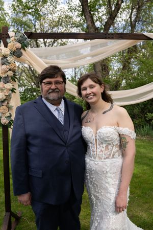 Bride in lace mermaid gown poses with older man in navy suit under floral arch.
