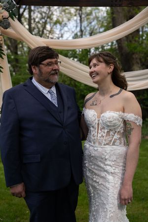 Bride in lace off-shoulder gown smiles at older man in navy suit during outdoor wedding ceremony.