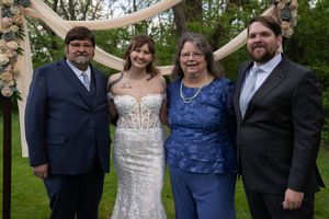 A bride in white corset gown poses with three family members under a floral arch at an outdoor wedding ceremony.