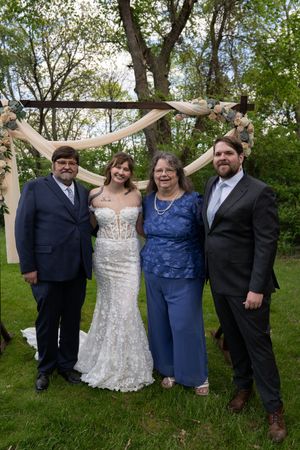 Wedding party of four posing under floral arch outdoors, bride in white lace gown with family in blue and navy attire.