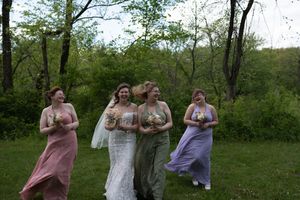 Bride in white lace gown laughing with four bridesmaids in colorful dresses outdoors in green wooded setting.
