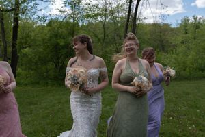 Smiling bride in strapless lace gown walking with bridesmaids in colorful dresses through green park.