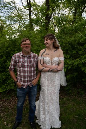 Bride in lace off-shoulder gown smiling with bearded man in plaid shirt outdoors.