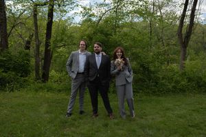 Three elegantly dressed wedding guests pose in gray suits on a green grassy woodland area.