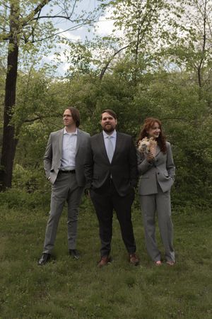 Groom with two groomsmen in gray suits standing in a lush green garden, one holding a small dog.