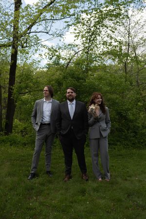 Three formally dressed wedding guests stand together on green grass surrounded by lush spring trees outdoors.