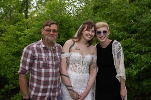 Bride in lace wedding gown poses with two guests in lush green garden setting.
