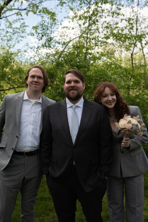 Three people posing outdoors at a wedding, two in gray suits and one in a dark suit holding a bouquet.