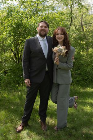 Groom in dark suit and bride in gray pantsuit holding bouquet pose in lush green outdoor setting.