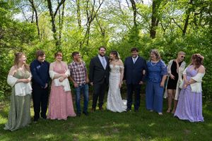 Wedding party group photo with bride and groom surrounded by family in lush garden.