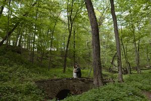 Bride and groom kissing on stone bridge surrounded by lush green forest.